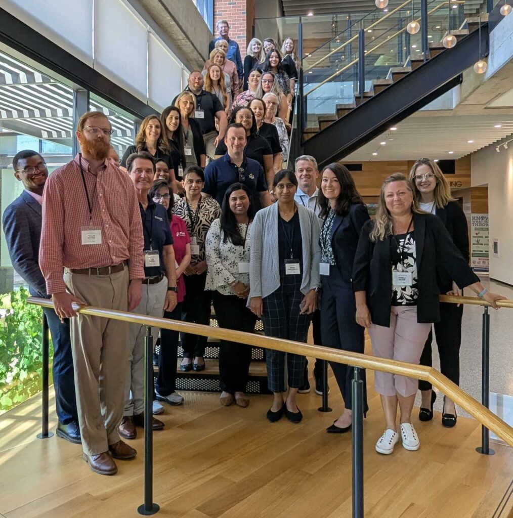 A group of smiling people on a set of stairs, wearing lanyards from their respective organizations.