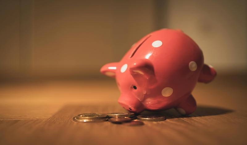 a pink piggy bank leaning over scattered coins on a wooden desk.